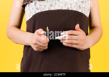 Primo piano di una donna incinta in pigiami marroni che tengono una sigaretta e un accendisigari su sfondo giallo. Stile di vita malsano durante la gravidanza. ni pericoloso Foto Stock