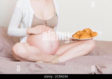 Primo piano di una giovane donna incinta ama mangiare croissant a letto. Pasticceria malsana durante il concetto di gravidanza. Foto Stock