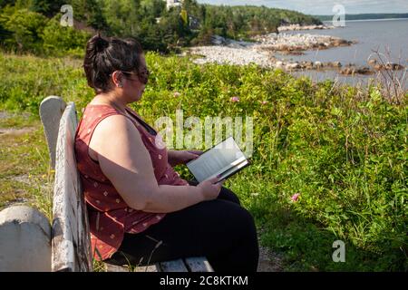 donna su una panchina tranquilla del parco accanto al mare che legge un buon libro al sole Foto Stock