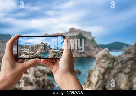 Il turista scatta foto della vista mozzafiato delle mura della città di Dubrovnik. Uomo che tiene il telefono e scattare foto delle pareti della fortezza, della costa rocciosa e del mare blu a Dubr Foto Stock