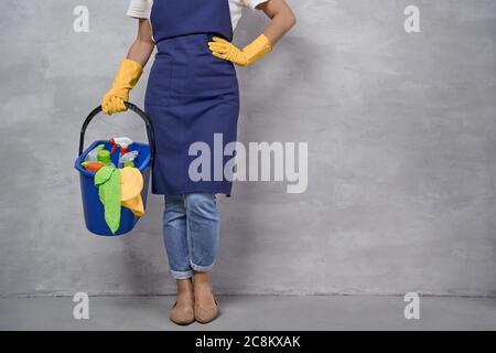 Ripresa di donna tagliata in guanti di gomma uniforme e gialla che tengono il secchio di plastica con stracci, detergenti e diversi prodotti per la pulizia mentre si trova contro la parete grigia. Servizi di pulizia, pulizie Foto Stock