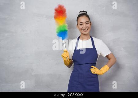 Concetto di servizio di pulizia e pulizia. Felice giovane donna, donna di pulizia in uniforme tenendo colorato pulitore in microfibra e sorridendo alla macchina fotografica mentre si trova contro parete grigia Foto Stock