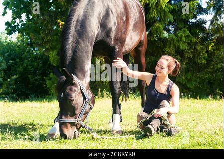 Giovane donna equestre seduta vicino al suo cavallo scuro su erba verde. Vivace immagine orizzontale in esterni multicolore in estate. Foto Stock