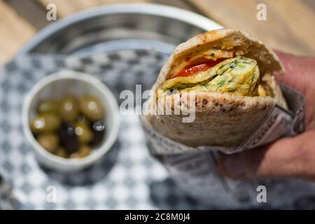 Mano dell'uomo che tiene un sandwich di omelette, in un pane pita, con pomodoro ed erbe, servito con olive Foto Stock