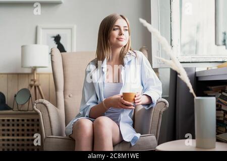 Bella giovane donna che beve caffè e guardando attraverso la finestra mentre si siede a casa in poltrona. Foto Stock