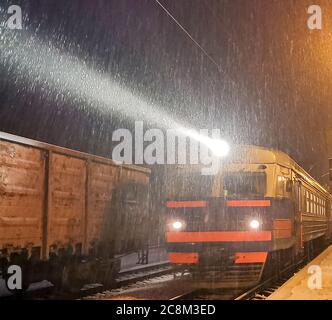 Sera treno arriva alla stazione. Nevicata complica il trasporto di passeggeri, blocchi di neve. Blizzard e derive di neve Foto Stock