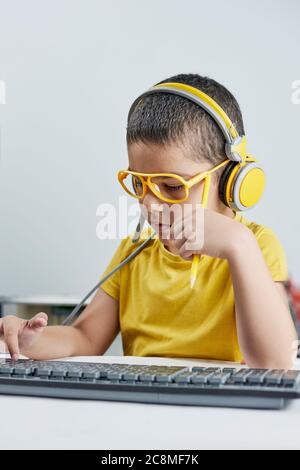 Un adorabile bambino in una t-shirt gialla con un corso online di ascolto delle cuffie. Ragazzo che scrive sulla tastiera del pc. Homeschooling. Carino ragazzo di razza mista Foto Stock