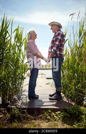 coppia senior che tiene le mani, guardandosi, sorridendo, in piedi su piattaforma di legno vicino al fiume Foto Stock