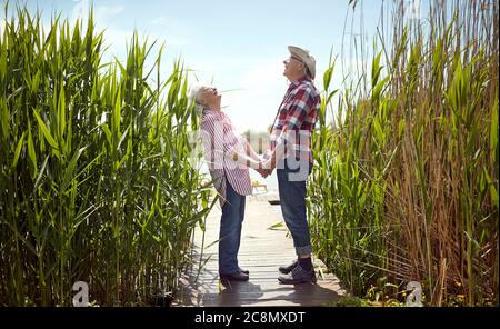 coppia senior che tiene le mani, guardando in alto, ridendo, in piedi su piattaforma di legno vicino al fiume Foto Stock