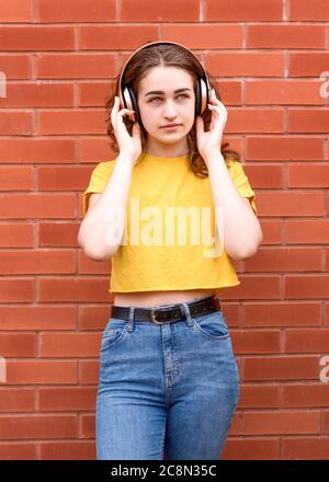 giovane donna che indossa una camicia gialla che ascolta la musica con le cuffie contro la parete in mattoni Foto Stock