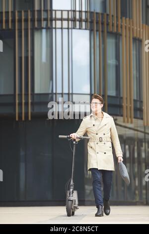 Ritratto verticale a lunghezza intera di gioioso giovane uomo d'affari che cammina accanto allo scooter elettrico mentre si viaggia per lavorare in città, spazio copia Foto Stock