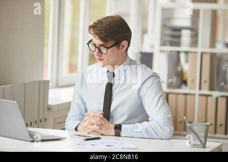 Ritratto di un giovane uomo d'affari che indossa occhiali seduti alla scrivania in ufficio e guardando lo schermo del computer portatile durante la videochiamata, spazio per la copia Foto Stock