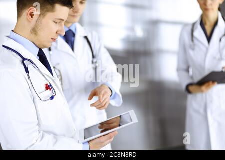 Gruppo di medici stanno controllando i nomi medici su un tablet per computer, con un infermiere con una clipboard sullo sfondo, in piedi insieme in un ospedale Foto Stock