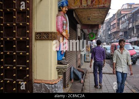 Pedoni che passano il piccolo Tempio Satyanarayano in Kika Street, Gulalwadi, Mumbai, India, una statua custode accanto al suo ingresso, un uomo che dormiva nelle vicinanze Foto Stock