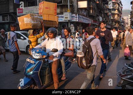 Un uomo guida la sua moto attraverso la trafficata Kalbadevi Road a Bhuleshwar, Mumbai, India, portieri passati con merci sulla testa e numerosi pedoni Foto Stock