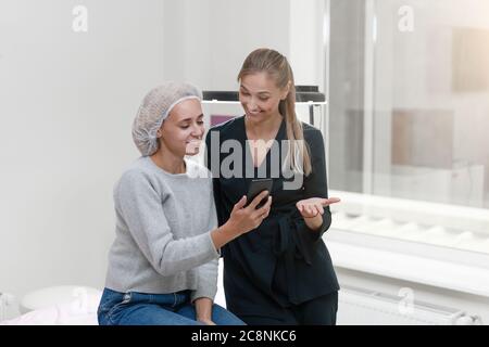 Armadio di cosmetologia client seduto sul divano. Gli stand estetista mostrano lo schermo dello smartphone e sorrisi. I pazienti con cosmetologo discutono delle opzioni di scelta del trattamento. Preparazione per la procedura di trucco permanente. Foto Stock