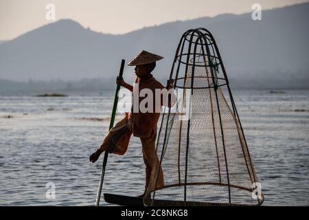 Lago Inle, Myanmar - Febbraio 2020: Tradizionale pescatore Intha gamba-rowing con una rete conica illuminata dal sole che tramonta. Foto Stock