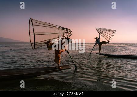 Lago Inle, Myanmar - Febbraio 2020: Tradizionali pescatori Intha gamba-rowing con reti coniche illuminate dal sole che tramonta. Foto Stock