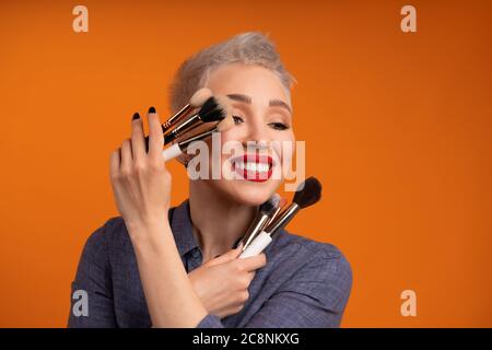 Primo piano artista trucco ritratto. Preparare i corsi. Concetto di autosevimento magistrale. Donna tenere le spazzole per il trucco sulle mani. Studio girato su sfondo arancione. Persone positive sorridenti Foto Stock