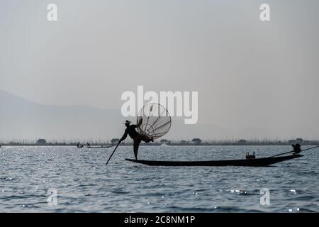 Famosi pescatori di inthe leg rowing sul lago Inle. Silhouette figura maschile in piedi su prua di barca lunga con sfondo paesaggio nebby Foto Stock