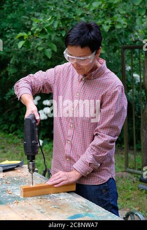 Craftswoman con occhiali di sicurezza che perfora un foro con una macchina elettrica in tavola di legno. Legno, fai da te, concetto di uguaglianza di genere. Foto Stock