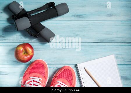Concetto di fitness, sneakers rosa, mela rossa, manubri e notebook con matita su sfondo in legno, vista dall'alto Foto Stock