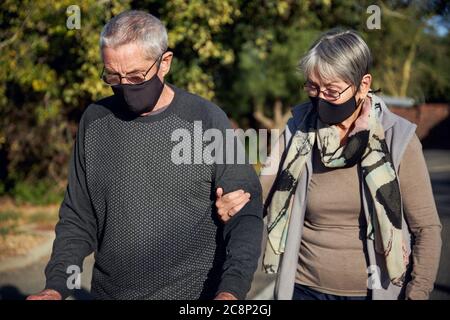 Coppia senior attiva sulla passeggiata all'aperto indossando maschere per il viso. Donna che aiuta l'uomo a camminare lungo la strada. Foto Stock