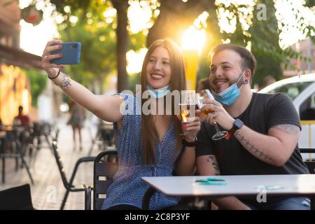 Spagna Madrid. Coppia caucasica tostatura con birra indossando maschera sanitaria prendendo un selfie. Riaprire pub e club dopo quarentine coronavirus Covid-19. M Foto Stock