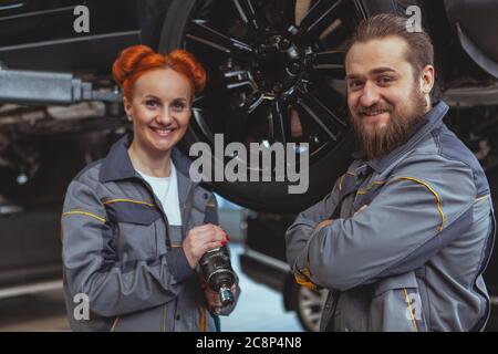 Allegro bella meccanica femminile e il suo collega maschio sorridente alla macchina fotografica, lavorando alla loro stazione di servizio auto. Rappresentante di tecnici di auto esperti Foto Stock