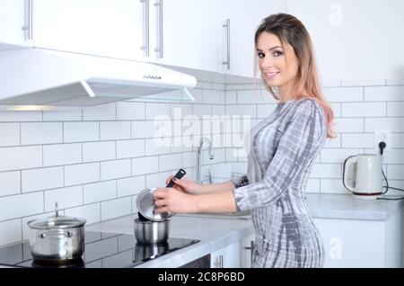 Giovane donna in abito grigio in cucina che prepara il pranzo in pentola. Bella casalinga dalla Polonia.. Foto Stock