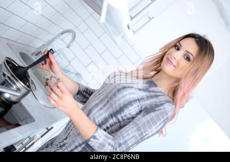 Giovane donna in abito grigio in cucina che prepara il pranzo in pentola. Bella casalinga dalla Polonia.. Foto Stock