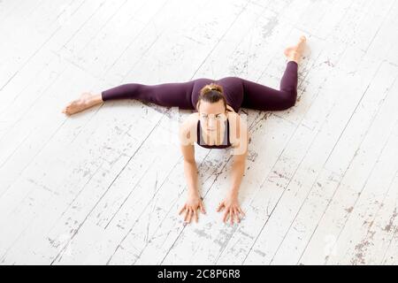 Vista ad alto angolo di una giovane donna atletica che fa una metà frontale split yoga posa su un pavimento bianco palestra con copyspace in uno stile di vita attivo e fitness Foto Stock