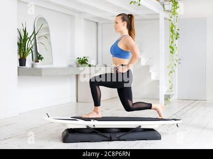 Giovane donna sportiva che fa un affondo statico posa su un surfset in una palestra high key con piante in un concetto di stile di vita sano Foto Stock