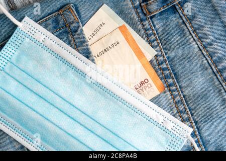 Primo piano di una maschera medica protettiva e banconote in euro nella tasca di vecchi jeans blu su sfondo grigio di legno. Crisi finanziaria globale e unem Foto Stock