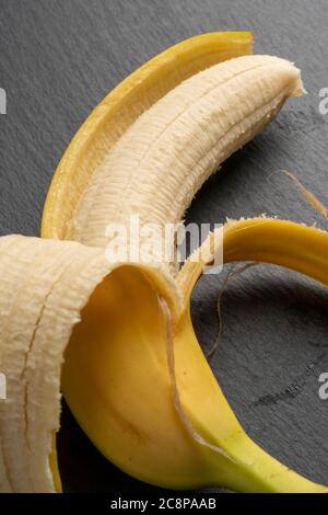 Banana gialla matura con bucce a metà buccia su una tavola scura di pietra grigia. Vista verticale. Concetto di dieta Foto Stock