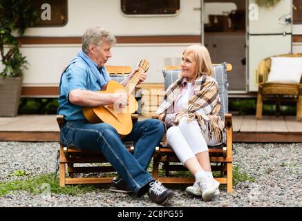 Uomo anziano romantico che suona la chitarra e canta la canzone alla moglie vicino a casa di guida al campeggio Foto Stock