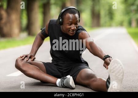 Sorridente sportivo nero seduto sul sentiero del parco e stretching Foto Stock
