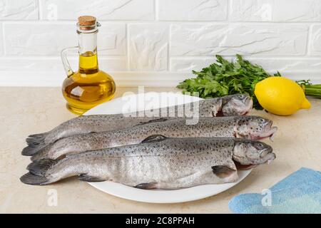 Tre pesci trota grezzi eviscerati su un piatto bianco su un tavolo da cucina preparato per cucinare. Piatti di pesce fresco, cibo sano e cucina a casa. Foto Stock