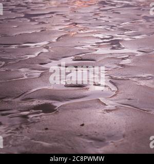 La spiaggia bagnata e le pozzanghere d'acqua illuminate da un sole che tramonta causando un soffuso bagliore rosa sulla sabbia Foto Stock