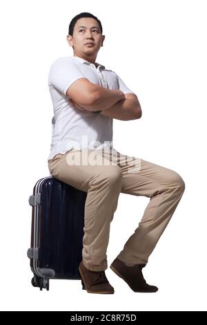 Uomo asiatico viaggiatore seduto sul suo bagaglio valigia, gesto d'attesa, indossare camicia casual e zaino, ritratto a lunghezza intera isolato su bianco Foto Stock