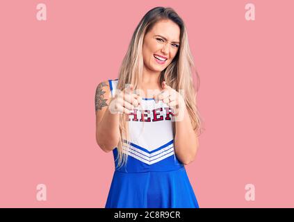 Giovane bella donna bionda con uniforme cheerleader puntando le dita alla macchina fotografica con viso felice e divertente. Buona energia e vibrazioni. Foto Stock