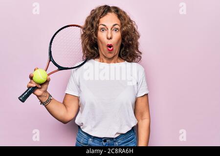 Media età bella sportiva giocando a tennis tenendo racchetta e palla su sfondo bianco spaventato e stupito con bocca aperta per sorpresa, sbelie Foto Stock