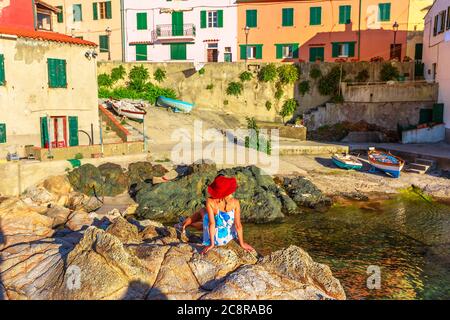 Donna turistica che riposa sulle scogliere di Marciana Marina a Borgo al Cotone, Isola d'Elba, Italia famose case di vecchio villaggio nella baia di Marciana Marina Foto Stock