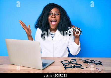 Bella donna africana che tiene occhiali da ottometria celebrando la vittoria con sorriso felice e vincitore espressione con mani sollevate Foto Stock