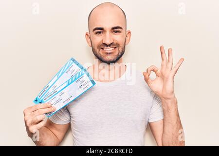 Giovane bel calvo uomo che tiene la carta d'imbarco della compagnia aerea facendo ok segno con le dita, sorridente amichevole gesturing simbolo eccellente Foto Stock