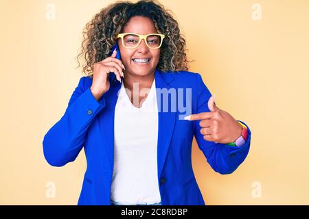 Giovane afroamericano più taglia donna che indossa occhiali che parlano sullo smartphone puntando il dito verso un auto sorridente felice e pro Foto Stock