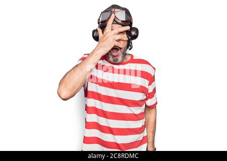 Aviatore di mezza età uomo che indossa casco vintage e occhiali su isolato sfondo bianco picchiando in shock coprendo viso e occhi con la mano, guardando th Foto Stock