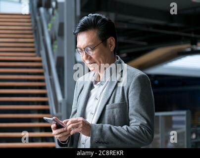 Uomo d'affari cinese che indossa una giacca, sorridendo e guardando il cellulare in mano. Foto Stock