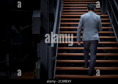 Vista posteriore di uomo d'affari con giacca a piedi su scala in legno. Foto Stock