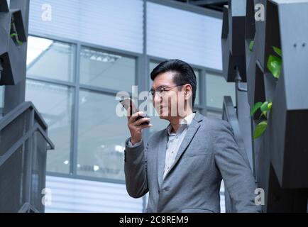 Uomo d'affari cinese che indossa una giacca, sorridendo e utilizzando il suo cellulare per lasciare un messaggio vocale Foto Stock